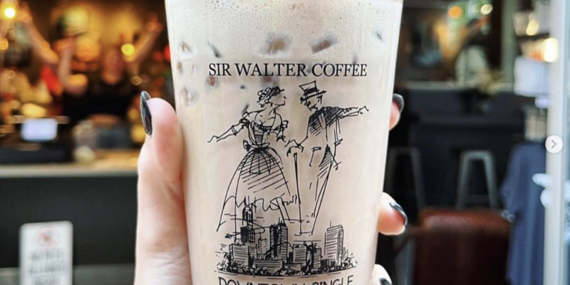 A hand holding a Sir Walter Raleigh coffee drink in front of the store